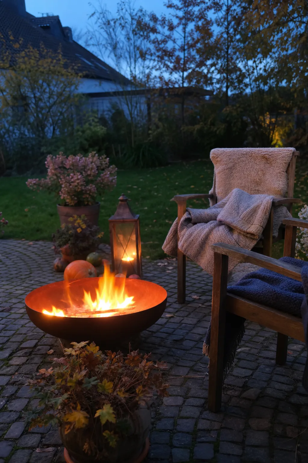Rostige Feuerschale mit Feuer auf einer Gartenterrasse, zwei Holzstühle mit Wolldecken, Laternen und Kürbisse daneben