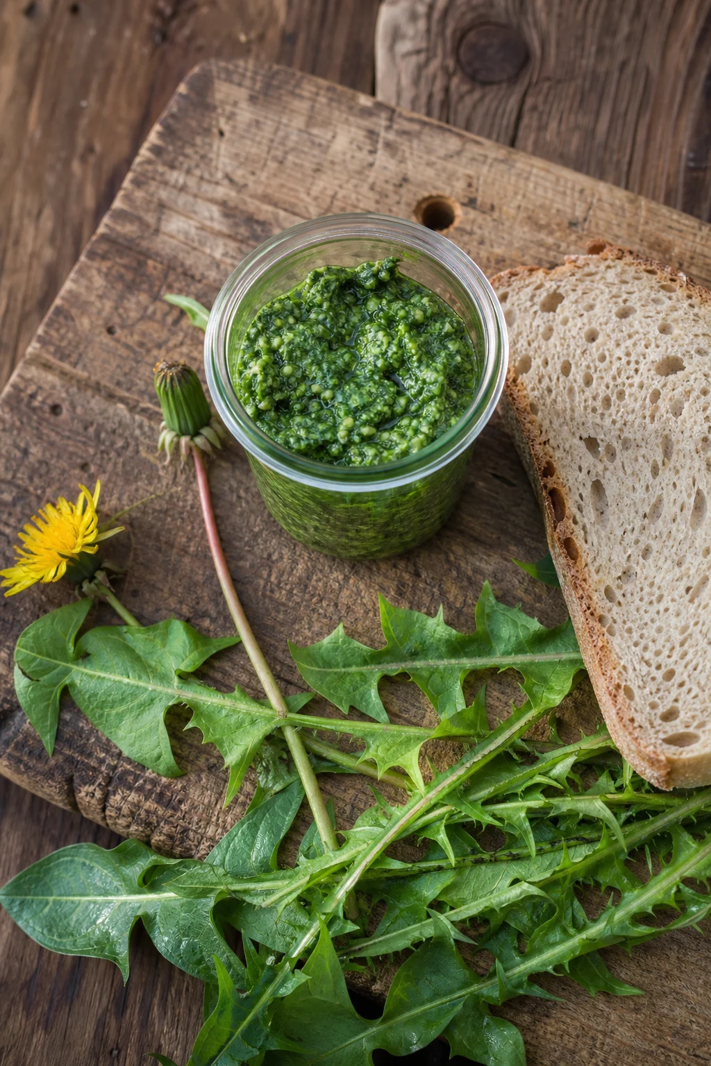 Kleines Glas mit leuchtend grünem Löwenzahn-Pesto auf rustikalem Holzbrett, daneben rohe Blätter und eine Scheibe Sauerteigbrot.