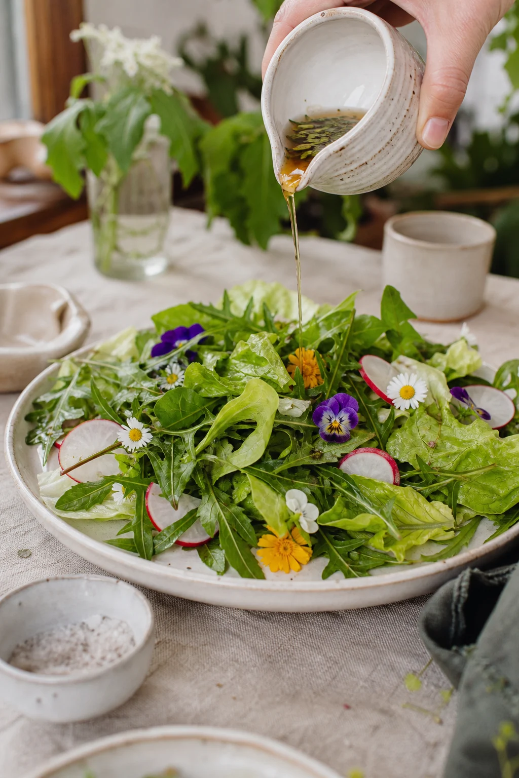 Frühlingssalat aus jungen Löwenzahnblättern, Pflücksalat, essbaren Blüten und Radieschen wird mit Wildkräuter-Essig aus einem Keramikkrug beträufelt.